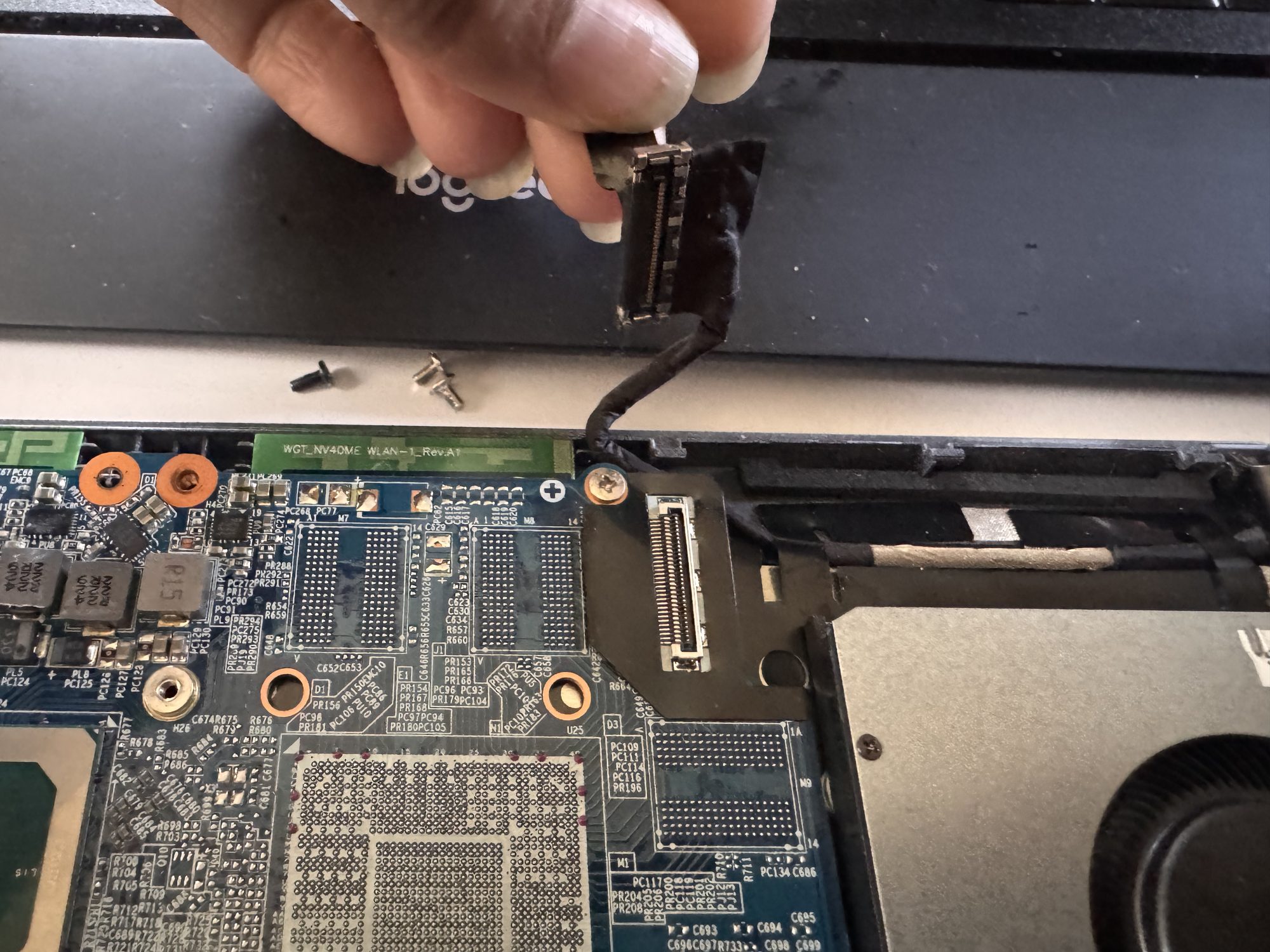 Close-up of a hand unplugging the thin ribbon cable LCD connector near the WLAN card, with two loose screws on the case beside it.